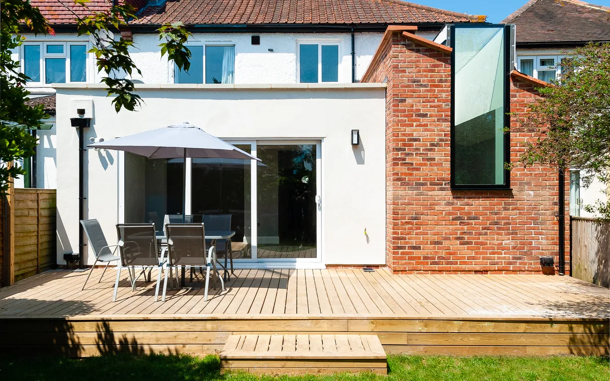 Rear extension with white render, a brick side wall with a tall slot window and sliding doors onto a timber deck
