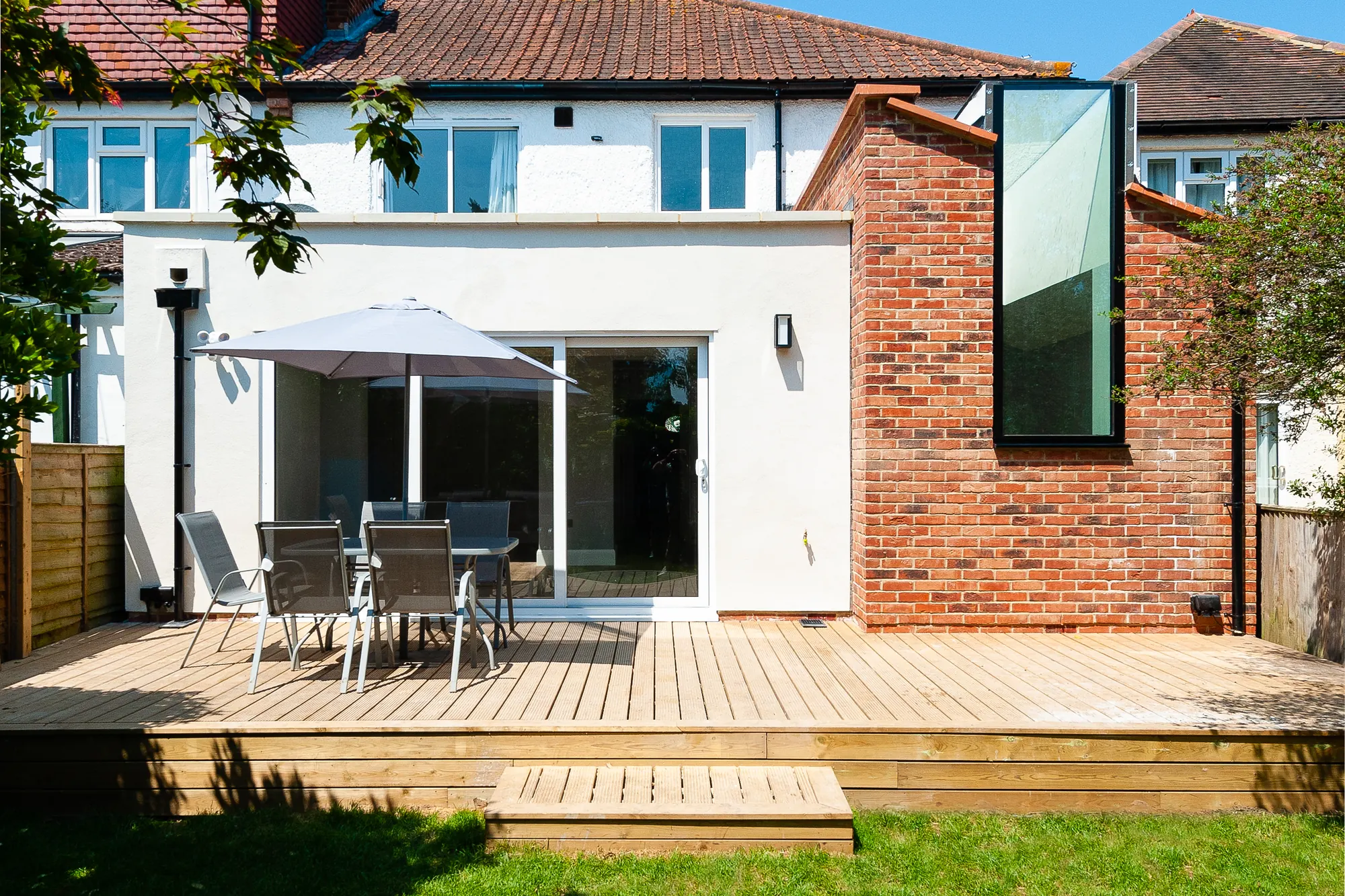 Rear extension with white render and a brick side wall with a tall glazed slot, opening onto a timber deck and lawn