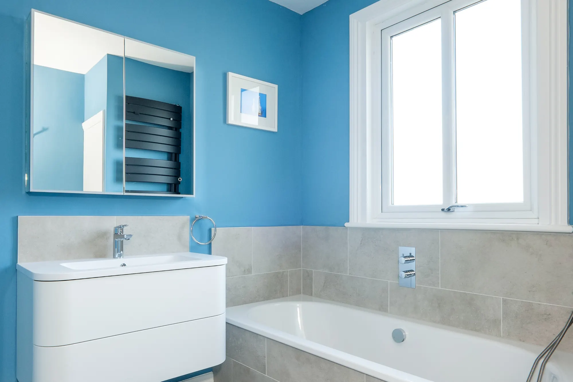Family bathroom with a white wall-hung vanity, a mirrored cabinet, stone-effect tiling and a built-in bath beneath the window