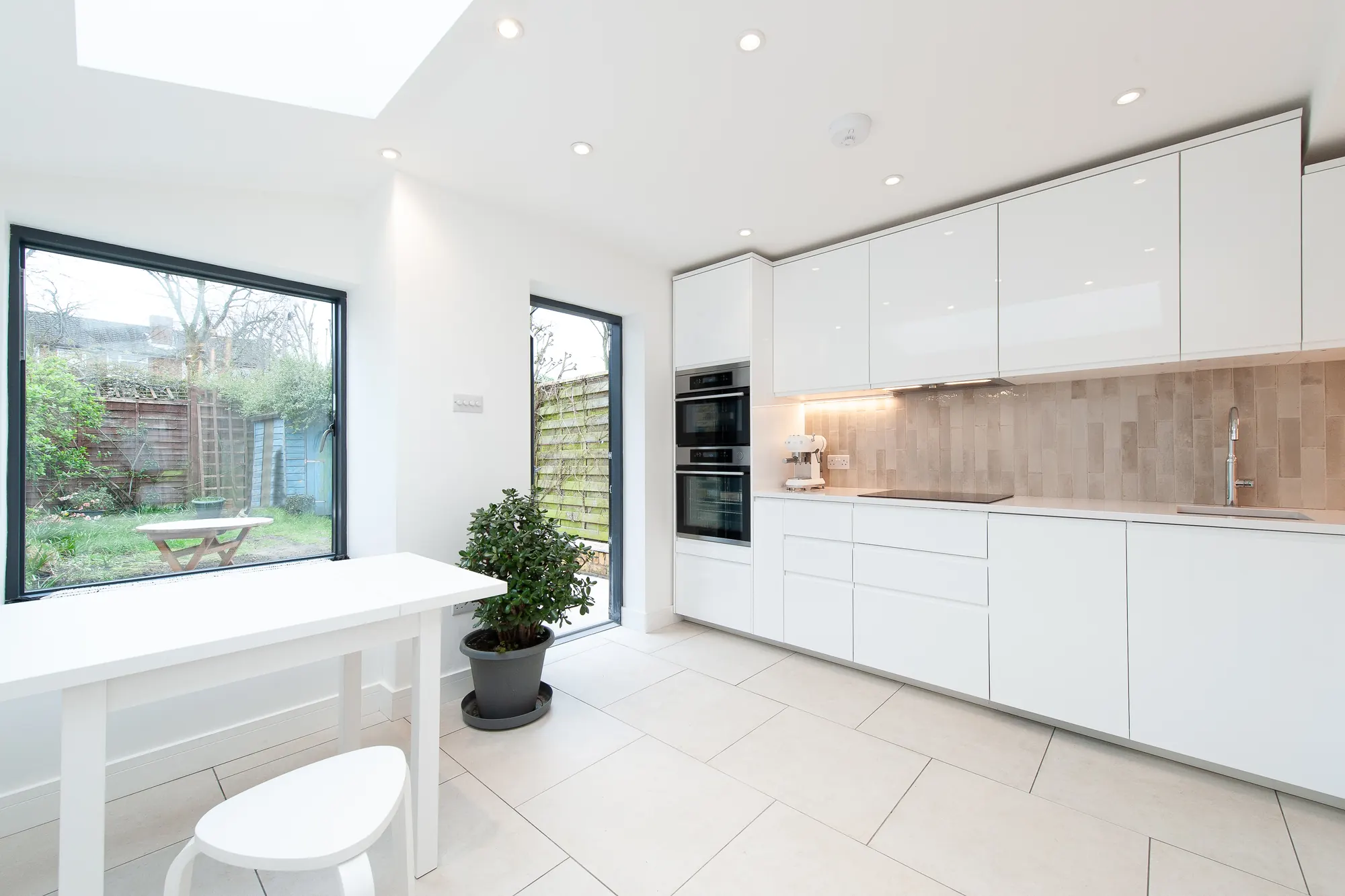 Bright white kitchen with a warm textured tile splashback, a small dining table and full-height glazing onto the garden