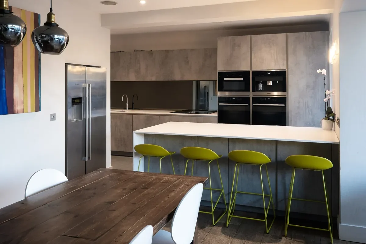 Completed kitchen: walnut dining table in front of a white island with lime green bar stools, concrete-effect units and tall ovens