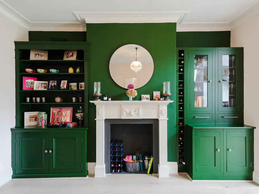 Deep green built-in shelving and cabinets either side of a white fireplace with a round mirror