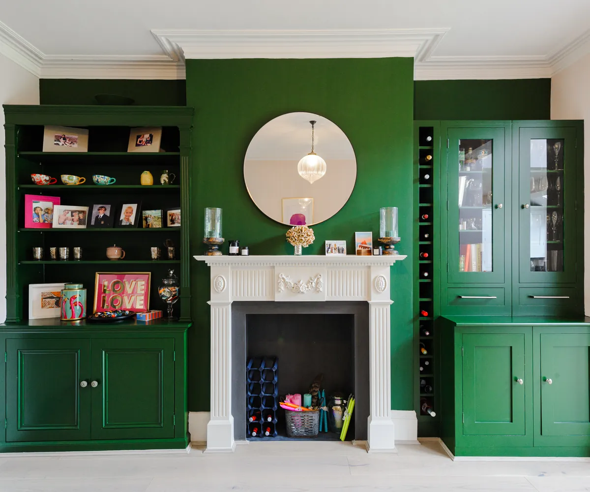 Deep green built-in shelving and cabinets either side of a white fireplace with a round mirror