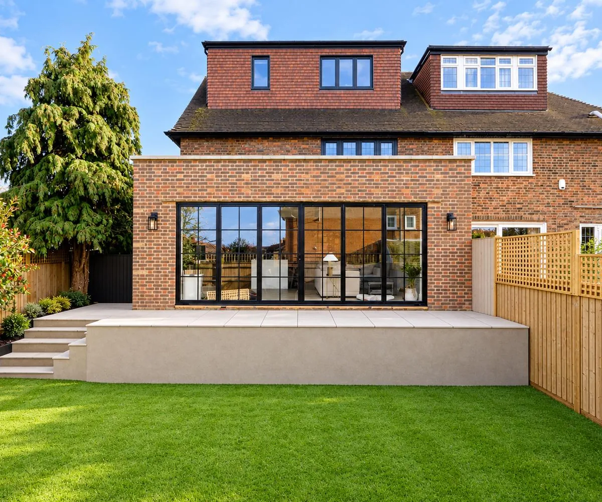 Brick rear extension with full-width black-framed glazing, seen across the garden lawn