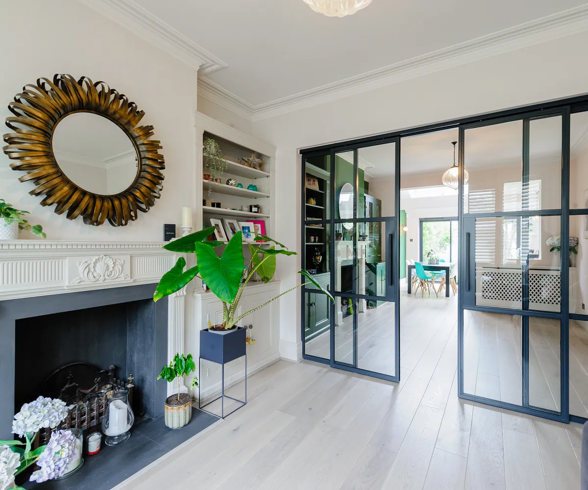 Period living room with a marble fireplace and sunburst mirror, opening through steel-framed glass doors